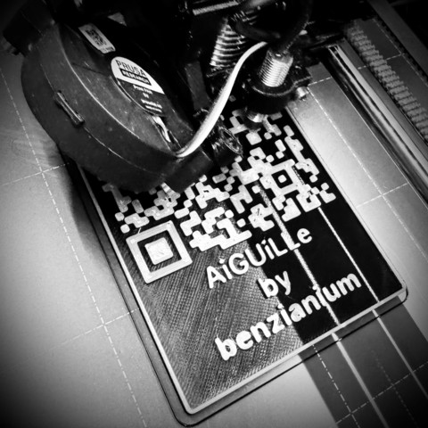 [GER] Schwarz/Weiss Nahaufnahme eines 3D-Drucker, der gerade eine Plakette mit einem QR-Code und der Aufschrift „AiGUiLLe by benzianium“ druckt.

[ENG] Close-up of a 3D printer head in the process of printing a black and white QR code with text "AiGuILLe by benzianium" on a flat surface.